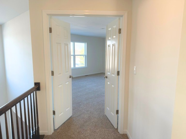 Spacious second-floor bedroom with double door entry, bright window, and modern neutral finishes in a two-story model home.
